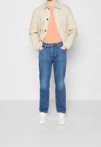 Giacca di cotone beige, t-shirt arancione, jeans in denim blu e sneakers bianche. L'outfit è casual con una vestibilità rilassata e dettagli minimi.