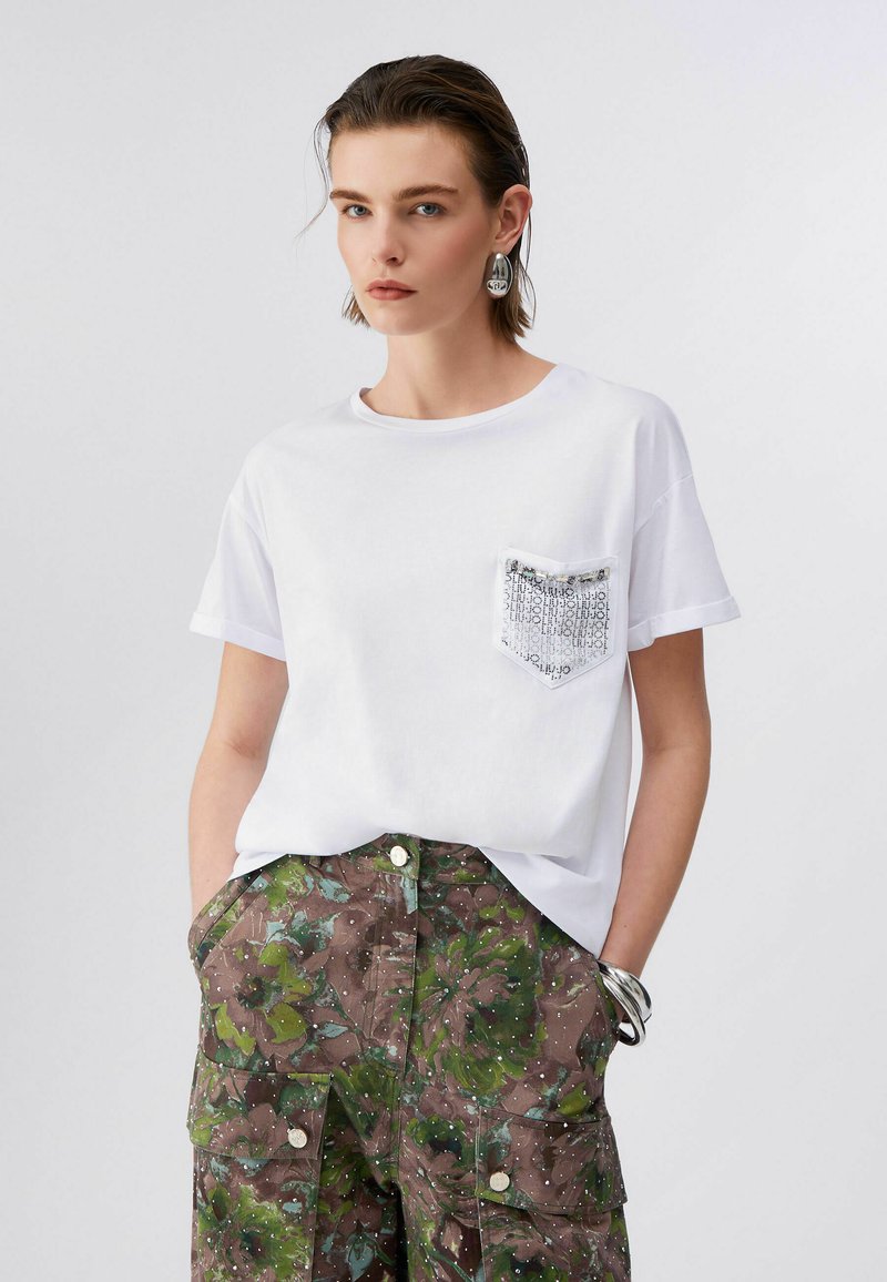 Jeune personne aux cheveux courts portant un t-shirt blanc avec une poche à motifs et un pantalon cargo vert à fleurs, posant les mains dans les poches.