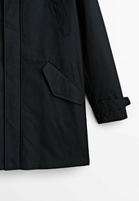 Manteau noir à manches longues avec poche à rabat inclinée et détail de sangle au niveau du poignet, présenté sur fond blanc.
