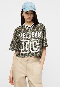 Νεαρή γυναίκα που φοράει μπλε καπέλο και κοντό πουκάμισο με λεοπάρ σχέδιο και κείμενο "ICECREAM IC", μπεζ παντελόνι ψηλόμεσο, ποζάροντας με το ένα χέρι στην τσέπη.