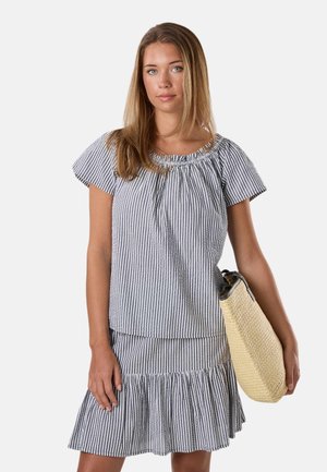 Femme portant un haut et une jupe à manches courtes rayés bleu et blanc, tenant un sac à main tissé beige sur son bras gauche.