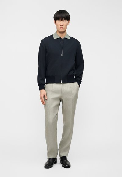 Ναυτικό μπουφάν τύπου bomber με ασημένιο φερμουάρ και πράσινο γιακά, συνδυασμένο με ανοιχτό γκρι παντελόνια και μαύρα παπούτσια. Λείο ύφασμα, άνετη εφαρμογή.