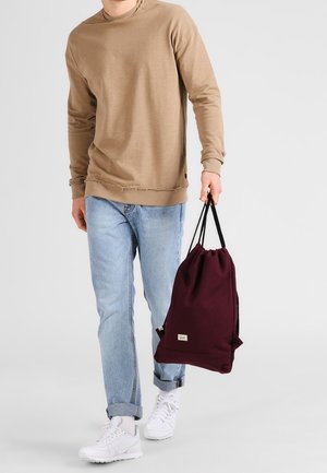 Borsa a tracolla bordeaux con tracolle nere, portata da una persona con una felpa beige e jeans azzurri, con orlo inferiore sfilacciato.