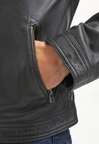 Hand teilweise in der seitlichen Reißverschlusstasche einer schwarzen Lederjacke mit sichtbaren Steppdetails.