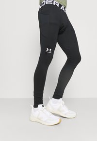 Svart, tettsittende treningslegginger laget av elastisk stoff, med et merket belte og logo på venstre ben. Satt sammen med hvite joggesko.