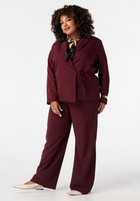 Costume bordeaux à deux pièces comprenant une veste à ceinture avec de larges manches et un pantalon large assorti. Écharpe noire et dorée à motif en accent.