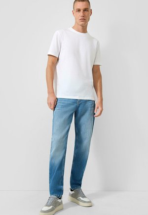 Mężczyzna ubrany w prostą białą koszulkę z krótkim rękawem, jasnoniebieskie dżinsy i szaro-białe trampki, stojący na tle jasnoszarego tła.