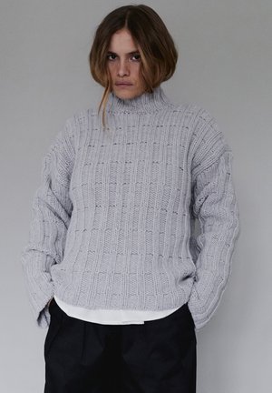 Szary sweter dziany z wysokim kołnierzem, z fakturowanym wzorem pionowych prążków. Luźny fason, noszony na białej koszuli i czarnych spodniach.