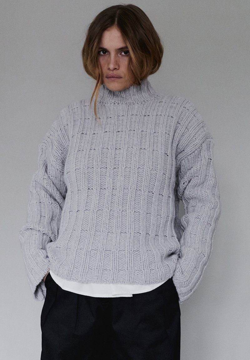 Szary sweter dziany z wysokim kołnierzem, z fakturowanym wzorem pionowych prążków. Luźny fason, noszony na białej koszuli i czarnych spodniach.
