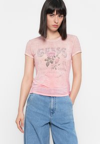 Femeie cu păr drept de culoare neagră, purtând un tricou grafic roz cu flori și blugi albastre cu talie înaltă, stând cu o mână în buzunar.