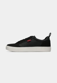 Tenis labă negru cu o suprafață texturată, având un model de logo, șireturi plate și o talpă din cauciuc albă, evidențiat de o etichetă de brand roșie.