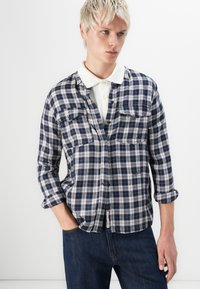 Camisa de cuadros en azul marino y blanco con cuello, mangas largas recogidas y dos bolsillos en el pecho; llevada sobre un polo blanco.