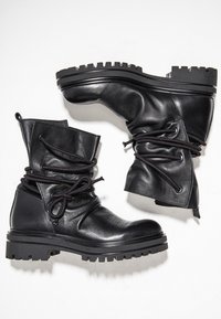 Botas de combate de cuero negro con suelas gruesas de tacos y cordones negros envueltos, mostradas una al lado de la otra sobre un fondo blanco.