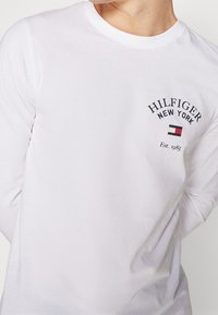 Weißes Langarm-T-Shirt aus Baumwolle, mit dem bedruckten Logo "HILFIGER NEW YORK" in Schwarz und einem kleinen farbigen Emblem auf der linken Brust.