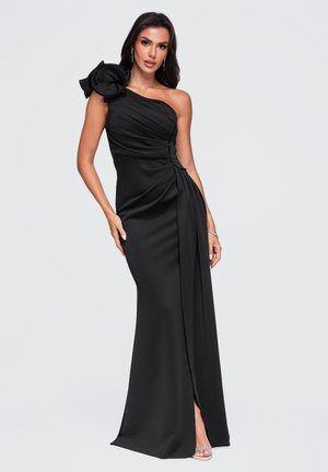 Un mannequin pose dans une robe noire longue jusqu’au sol, à une seule épaule, avec un grand détail floral sur l’épaule et une fente devant.