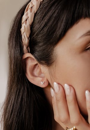 Silberne, schmetterlingsförmige Ohrringe mit klaren Edelsteinen, getragen von einer Person mit braunem Haar, einem geflochtenen rosa Haarband und neutralem Nagellack.