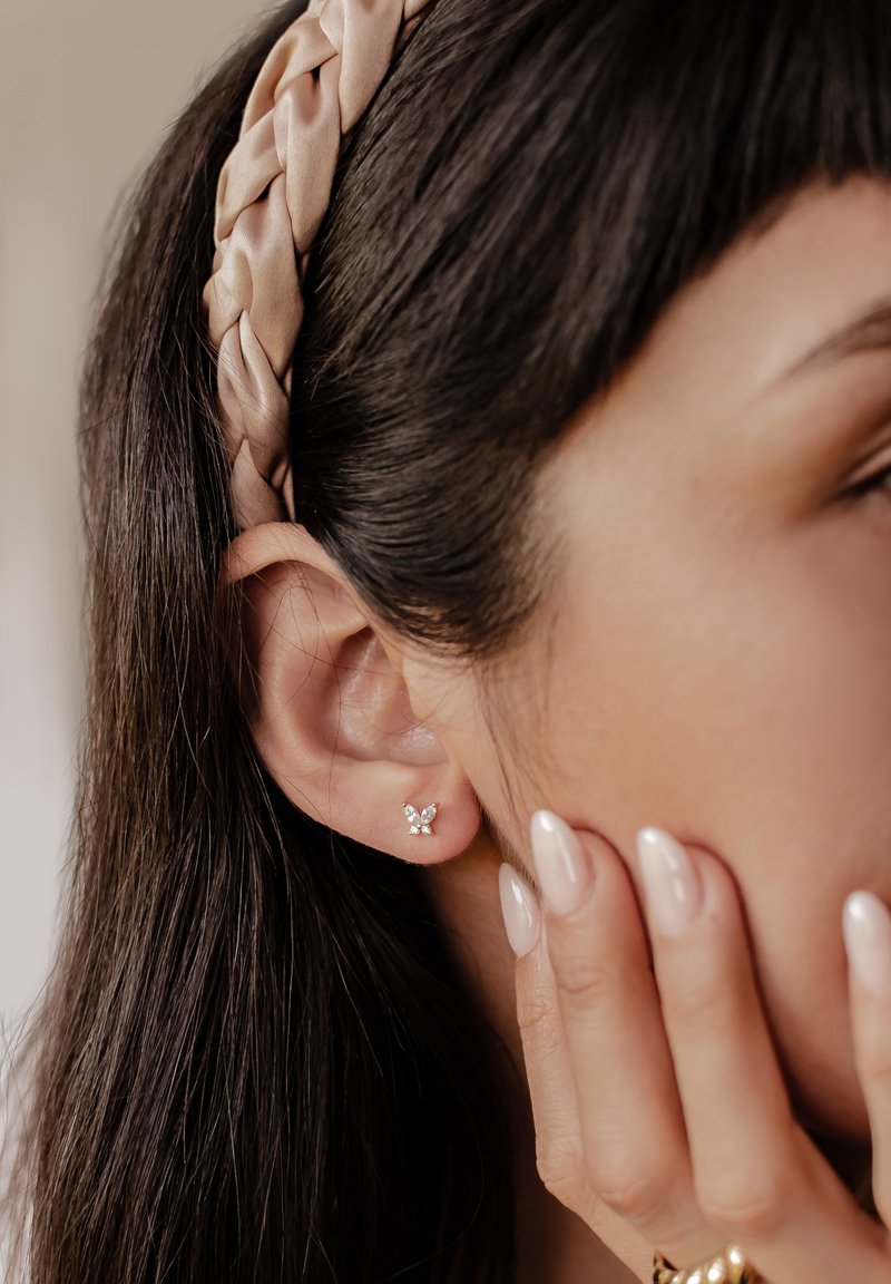 Boucles d'oreilles en argent en forme de papillon avec des pierres précieuses transparentes, portées par une personne aux cheveux bruns, avec un bandeau tressé rose et des ongles au vernis neutre.