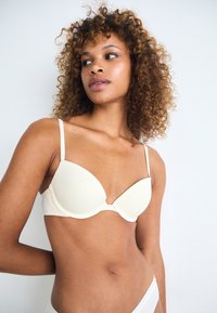 Beige polstret bh med glatte kopper, justerbare stropper og delikat blonderdetalj på båndet, komplementert av en nøytral bakgrunn.