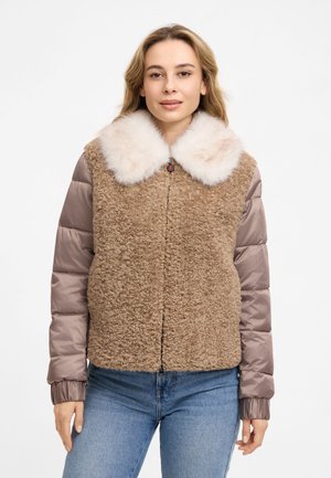 Frau trägt eine braune Sherpa-Jacke mit wattierten taupefarbenen Ärmeln und einem großen weißen Pelzkragen, kombiniert mit blauen Jeans.