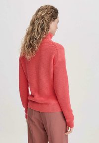 Korallenfarbener Strickpullover mit hohem Kragen, gerippten Bündchen und Saum, strukturiertem Muster und lässigem Schnitt, kombiniert mit hellrosa Hosen.