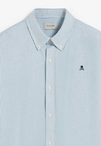 Camisa de botones a rayas azul claro y blanca con cuello. Presenta un pequeño logo de calavera y huesos cruzados en el lado izquierdo del pecho.