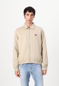 Beige zip-up jakke med spiss krage, elastiske mansjetter og kant. Har en liten brodert logo på venstre bryst.