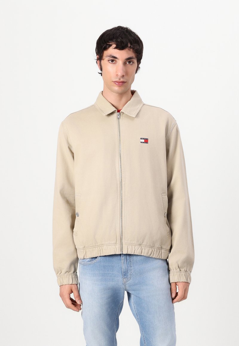 Beige zip-up jakke med spiss krage, elastiske mansjetter og kant. Har en liten brodert logo på venstre bryst.