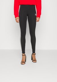 Svarta leggings med vita sidostråk, tillverkade av stretchigt material. Bärs med en röd sweatshirt och svarta klacksandaler. Minimalistisk Adidas-logotypdetalj.