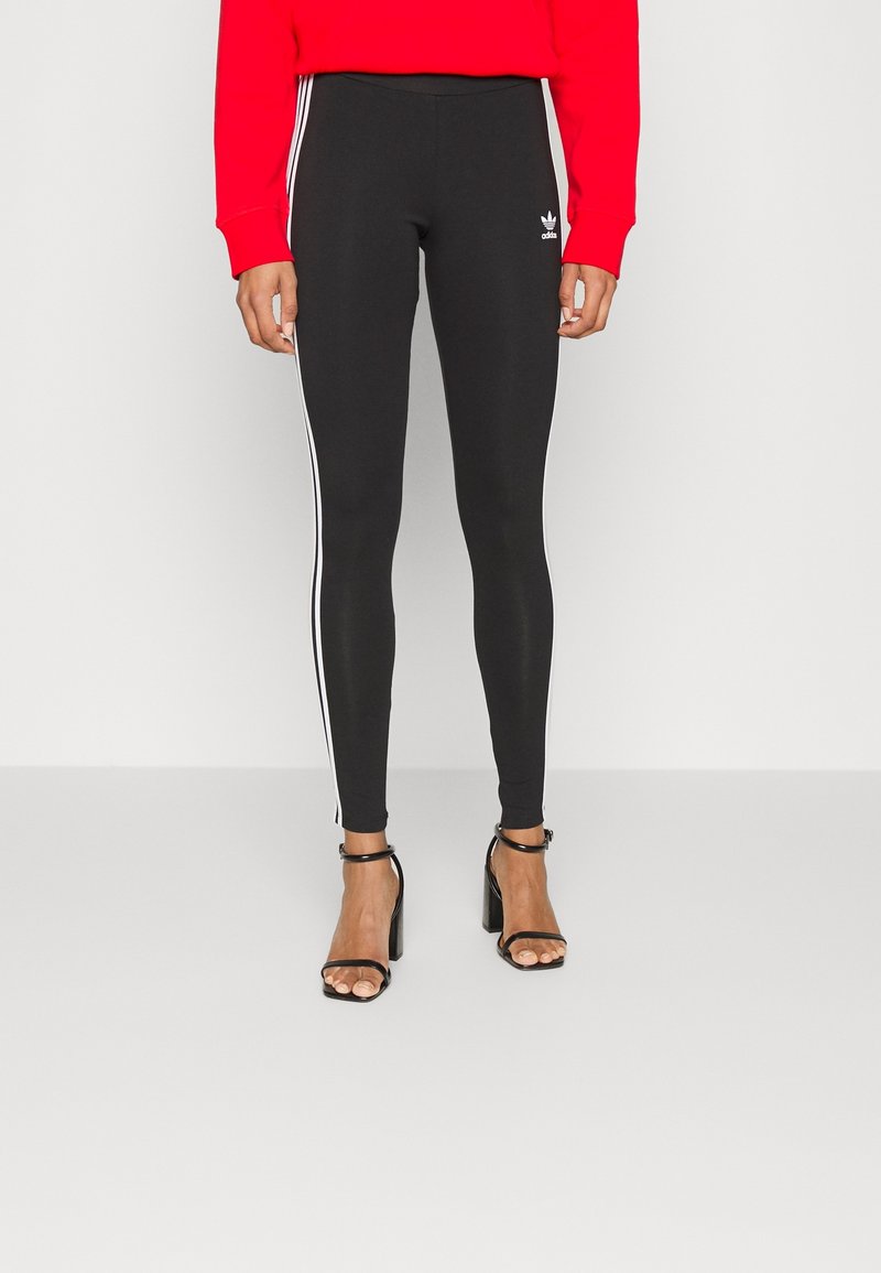 Svarta leggings med vita sidostråk, tillverkade av stretchigt material. Bärs med en röd sweatshirt och svarta klacksandaler. Minimalistisk Adidas-logotypdetalj.