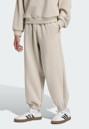 Beige joggerbukser med elastiske manchetter, afslappet pasform og sidelommer. Kombineret med hvide sneakers med sorte striber og brune detaljer.