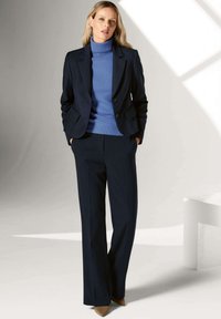 Marineblauwe op maat gemaakte blazer over een lichtblauwe coltrui, gecombineerd met donkere broeken en beige hakken. Glad materiaal en gestructureerd ontwerp.