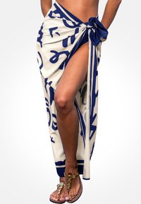 Weißer Sarong mit auffälligen blauen abstrakten Mustern, an der Taille gebunden; hat einen hohen Schlitz und kontrastierenden blauen Besatz. Goldene Sandalen sind sichtbar.