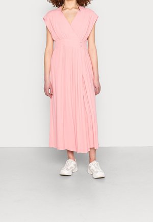 Frau trägt ein ärmelloses rosa plissiertes Midikleid mit V-Ausschnitt und weißen Sneakers, steht vor einem schlichten hellen Hintergrund.