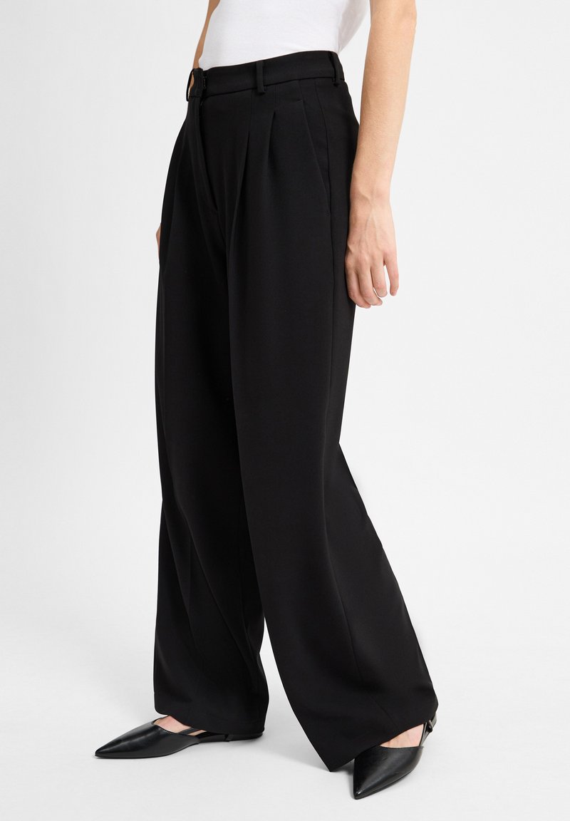 Zwarte wijde broek met plooien, op maat gemaakte tailleband en riemlussen. Gepaarde met puntige zwarte flats. Eenvoudig ontwerp, gladde textuur.