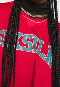 Rotes Netz-Jersey mit blauer und weißer Schrift "SILK" über der Brust, kombiniert mit geschichteten goldenen Halsketten. Strukturiertem Stoff und Details am Halsauschnitt.
