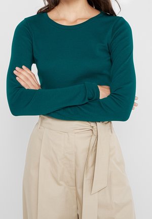 Personne les bras croisés portant un haut à manches longues bleu sarcelle et un pantalon beige taille haute avec une ceinture nouée.