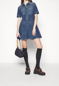 Vestido de denim con mangas cortas, botón en el frente y dos bolsillos en el pecho. Combinado con calcetas de canalé hasta la rodilla y zapatos de plataforma, sosteniendo un bolso pequeño.