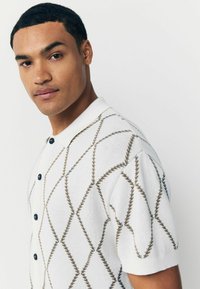 Weißes Poloshirt mit kurzen Ärmeln, das ein rautenförmiges Muster in Braun zeigt, mit einer Knopfleiste mit drei Knöpfen und strukturiertem Strickstoff.