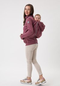 Felpa con cappuccio color mauve con pannello posteriore con zip per il trasporto del bambino. Modello abbinato a jeans skinny beige chiaro e sneakers pastello.