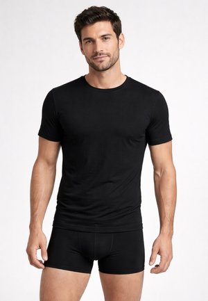 Uomo in piedi che indossa una maglietta nera aderente a maniche corte e boxer neri contro uno sfondo bianco uniforme.