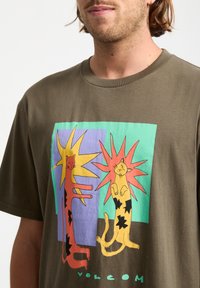 Oliven grøn bomulds T-shirt med et grafisk motiv af to farverige katte med stjernebaggrunde i rød, gul, blå og grøn. Tekst: "VOLCOM."