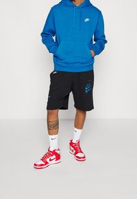 Man med blå Nike hoodie, svarta Nike shorts, vita Nike strumpor och röda och vita Nike sneakers, med en tatuering på sin högra vad.