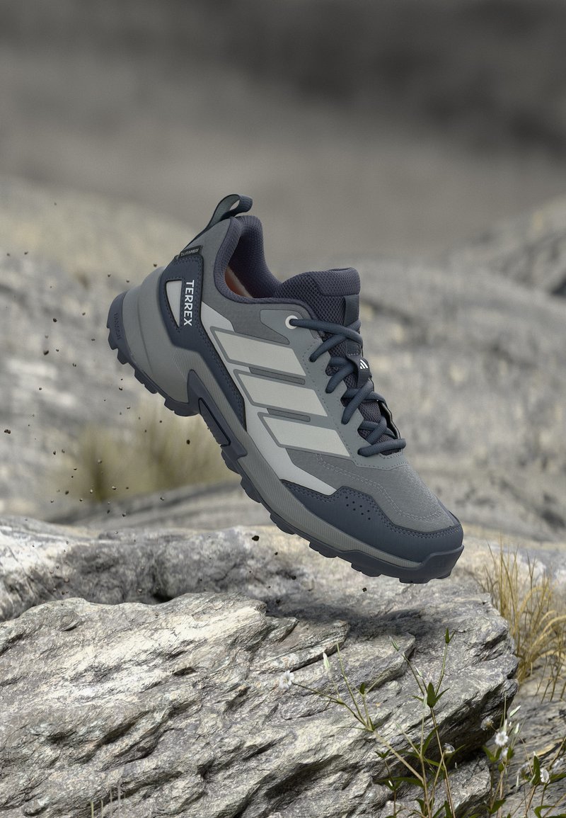 Grau-schwarzer Adidas Terrex Wanderschuh in der Luft über felsigem Gelände mit kleinen Pflanzen, der die robuste Sohle und das Outdoor-Design hervorhebt.