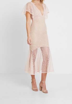 Mujer con un vestido midi de encaje rosa claro con mangas volanté y sandalias nude de tacón cuadrado con tira en el tobillo sobre un fondo blanco liso.