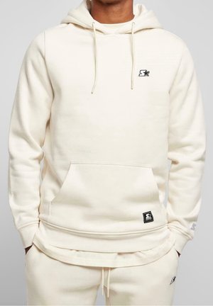 Mężczyzna w kremowej bluzie z kapturem i dopasowanych spodniach dresowych z małymi czarnymi naszywkami z logo, ręce w kieszeniach, stojący na jednolitym tle.