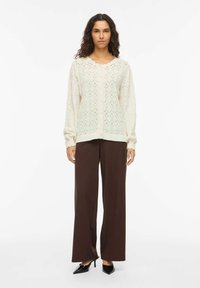Femme portant un cardigan blanc en tricot à boutons, un pantalon large marron et des chaussures à talons noires à bout pointu, debout devant un fond blanc uni.