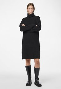 Femeie cu brațele încrucișate, purtând o rochie pulover cu guler înalt neagră și cizme negre de piele până la genunchi, stând în picioare pe un fundal alb simplu.