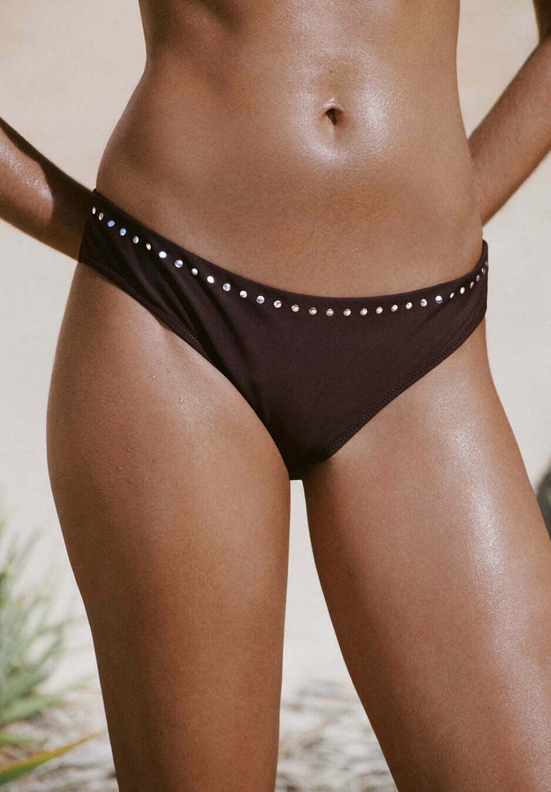 Milieu du corps d’une femme portant un bas de bikini noir avec de petits clous argentés le long de la ceinture, debout en plein air.