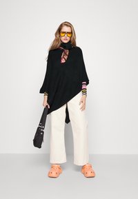 Μαύρο πόντσο με ψηλό λαιμό, με ριγέ μανίκια σε ροζ και κίτρινο. Συνδυασμένο με κρεμ παντελόνι σε φαρδιά γραμμή και πορτοκαλί γόβες. Μαύρη τσάντα αξεσουάρ.