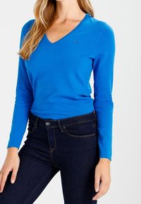 Sweter z dekoltem w serek, z długimi rękawami w jasnoniebieskim dzianinowym materiale; posiada mały akcent w postaci logo na piersi, zestawiony z ciemnoniebieskimi dżinsami.
